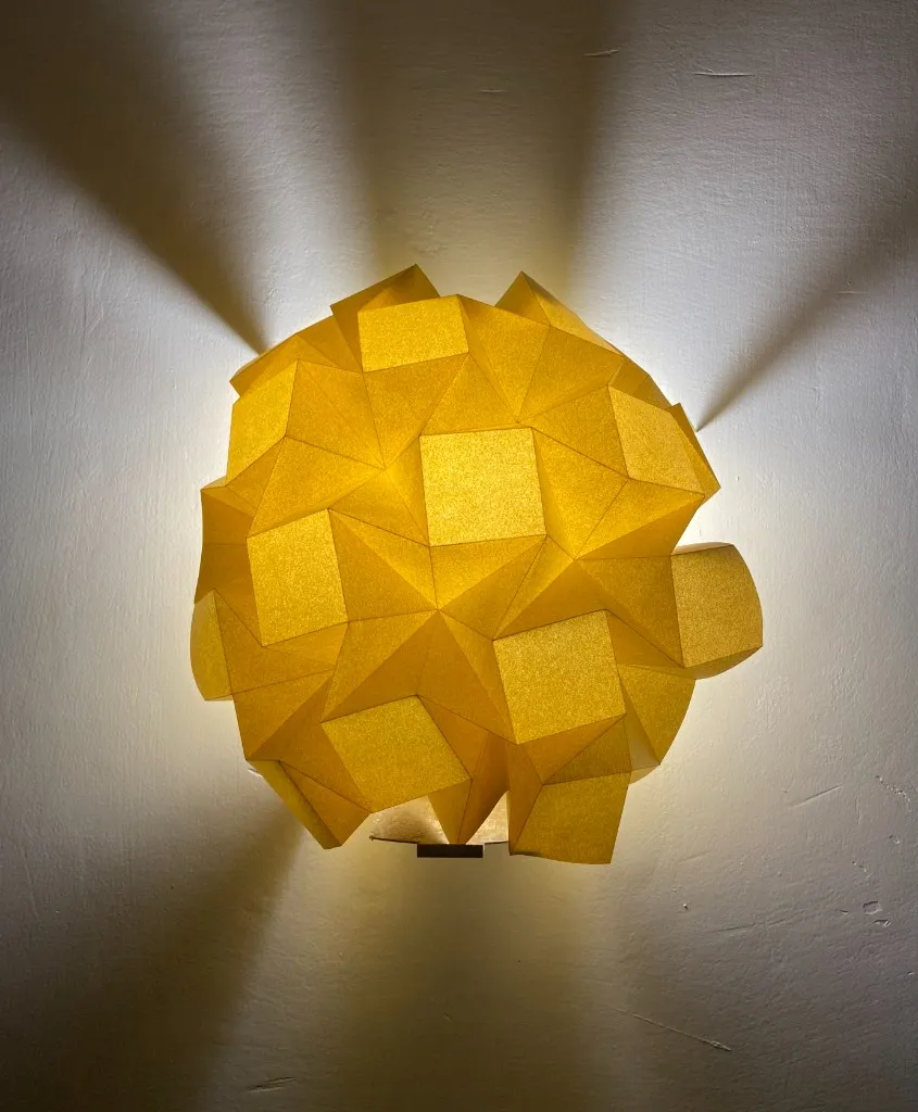 Glowing yellow modular origami sphere casting light on a wall